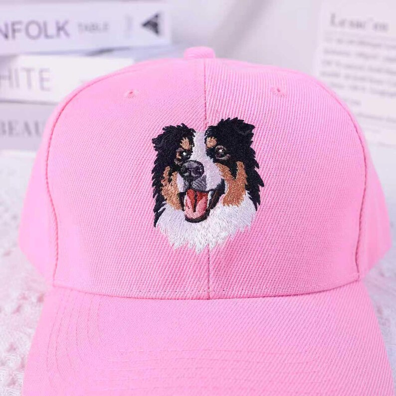 May include: A pink baseball cap featuring an embroidered image of a dog's face. The dog has black, brown, and white fur, with its mouth open. The cap is made of a soft fabric and has a curved brim.