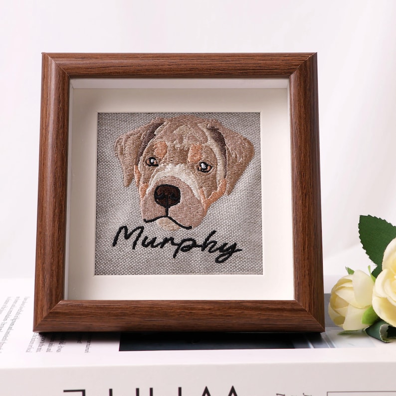 May include: A framed embroidery featuring a dog's face with the name "Murphy" stitched below. The dog's face is brown and tan, set against a gray background. The frame is a dark brown wood.