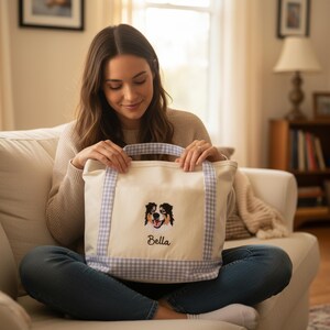 May include: A cream-colored canvas tote bag featuring light blue and white gingham patterned handles and a bottom trim. The bag displays an embroidered portrait of a tricolor dog above the name Bella stitched in a dark cursive font.