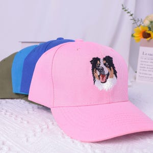 May include: A row of baseball caps in various colors: olive green, light blue, dark blue, and pink. The pink cap features an embroidered dog face with black, brown, and white details.