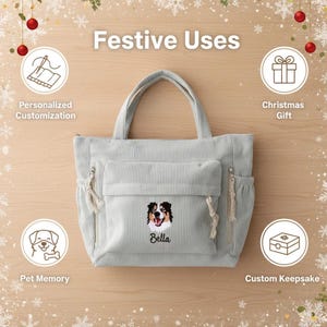 May include: A light grey corduroy tote bag featuring an embroidered portrait of a dog with the name Bella stitched below. The bag includes a large front pocket, two side pockets with rope-style zipper pulls, and two top handles for carrying.