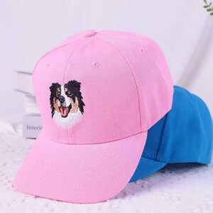 May include: A pink baseball cap with an embroidered dog face. The dog has black, brown, and white fur, with its tongue sticking out. A blue baseball cap is partially visible behind the pink cap.
