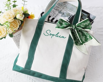 May include: A white canvas tote bag with green trim, handles, and the name "Sophia" embroidered in green. A green and white patterned scarf is tied to the handle. The bag is filled with magazines and a bouquet of flowers.