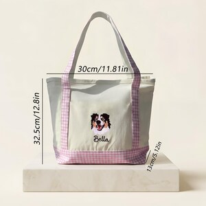 May include: A cream-colored canvas tote bag with light purple gingham handles and base. The bag features an embroidered dog face and the name Bella. Dimensions are 11.81 inches wide, 12.8 inches high, and 5.12 inches deep as indicated by text.