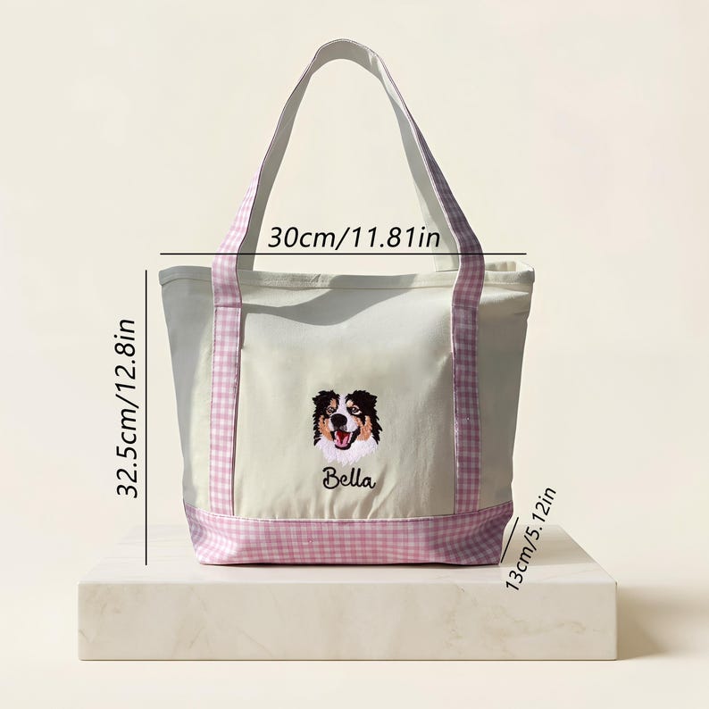 May include: A cream-colored canvas tote bag with light purple gingham handles and base. The bag features an embroidered dog face and the name Bella. Dimensions are 11.81 inches wide, 12.8 inches high, and 5.12 inches deep as indicated by text.