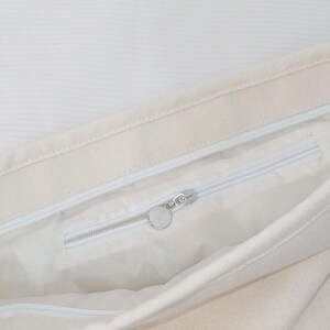 May include: An off-white canvas tote bag featuring a top zipper closure and an interior zippered pocket. The interior pocket includes a silver-toned circular zipper pull, providing a secure compartment within the main storage area of the bag.