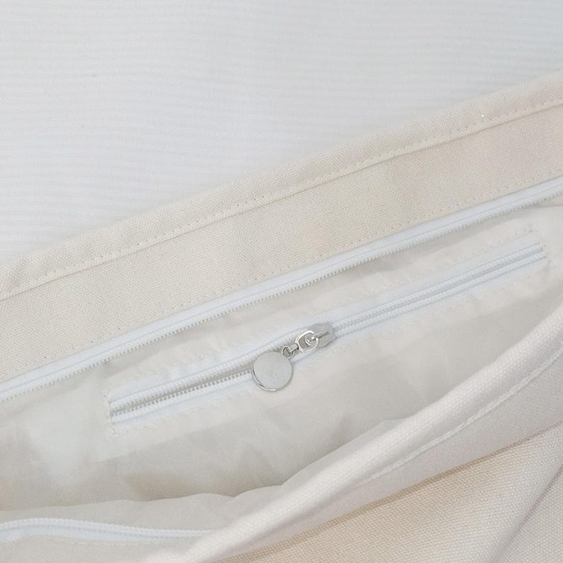 May include: An off-white canvas tote bag featuring a top zipper closure and an interior zippered pocket. The interior pocket includes a silver-toned circular zipper pull, providing a secure compartment within the main storage area of the bag.