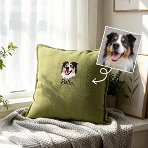 May include: A square olive green throw pillow with a textured herringbone pattern. The centre features an embroidered portrait of a black, white, and tan dog with the name Bella stitched in black cursive lettering below the dog's face.