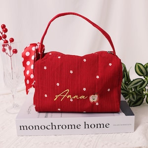 May include: A red handbag with a handle and a white polka dot ribbon. The bag is embroidered with white flowers and the name "Anna" in gold script, along with an apple design. The bag is sitting on a white book.