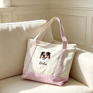 May include: A cream-colored canvas tote bag featuring a pink and white gingham pattern on the base and handles. The front displays an embroidered portrait of a tri-color dog with the name Bella stitched in black cursive lettering below the image.