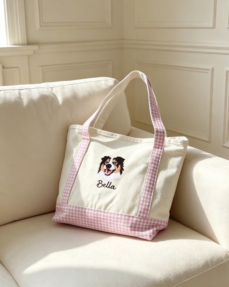 May include: A cream-colored canvas tote bag featuring a pink and white gingham pattern on the base and handles. The front displays an embroidered portrait of a tri-color dog with the name Bella stitched in black cursive lettering below the image.