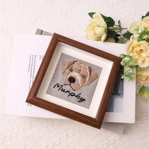 May include: A framed embroidered artwork featuring a dog's face with the name "Murphy" stitched below. The artwork is in a brown wooden frame, set against a neutral background with flowers.