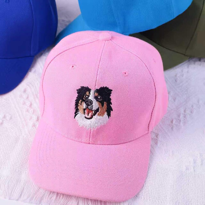 May include: A pink baseball cap featuring an embroidered dog face with black, brown, and white details. The cap has a curved brim and a structured crown. Other caps in blue and green are visible in the background.