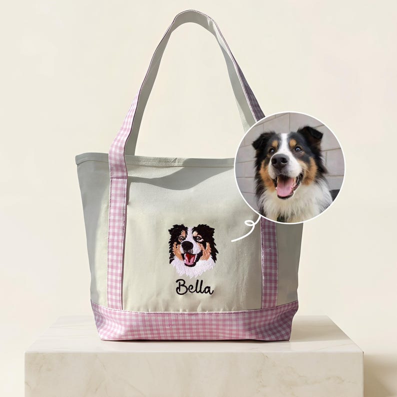 May include: A cream-colored canvas tote bag featuring a light purple and white gingham trim on the handles and base. The front displays an embroidered portrait of a tricolor dog above the name Bella in black script, with a circular photo inset.