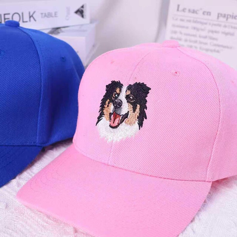 May include: A pink baseball cap with a curved brim and an embroidered image of a dog's face. The dog has black, brown, and white fur, with its tongue sticking out. A blue baseball cap is partially visible in the background.