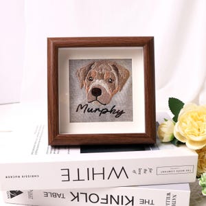May include: A framed embroidered portrait of a dog with the name "Murphy" stitched below. The dog's face is detailed with brown and white thread, set against a gray background. The frame is a dark brown color.