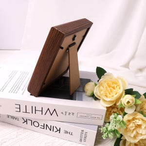 May include: A brown wooden picture frame with a stand, resting on a stack of books. The frame has a dark wood grain finish. Yellow roses and greenery are in the background.