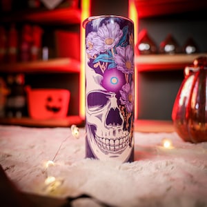 May include: A cylindrical tumbler with a skull design and floral accents. The tumbler features a purple background with a white skull and purple flowers. A glowing light is in the center of the skull. The tumbler is a unique gift for Halloween.