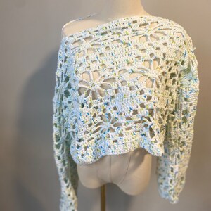 May include: A white, off-the-shoulder crochet top with long sleeves. The open weave design features a floral pattern and is speckled with blue and yellow threads. The top is displayed on a mannequin.