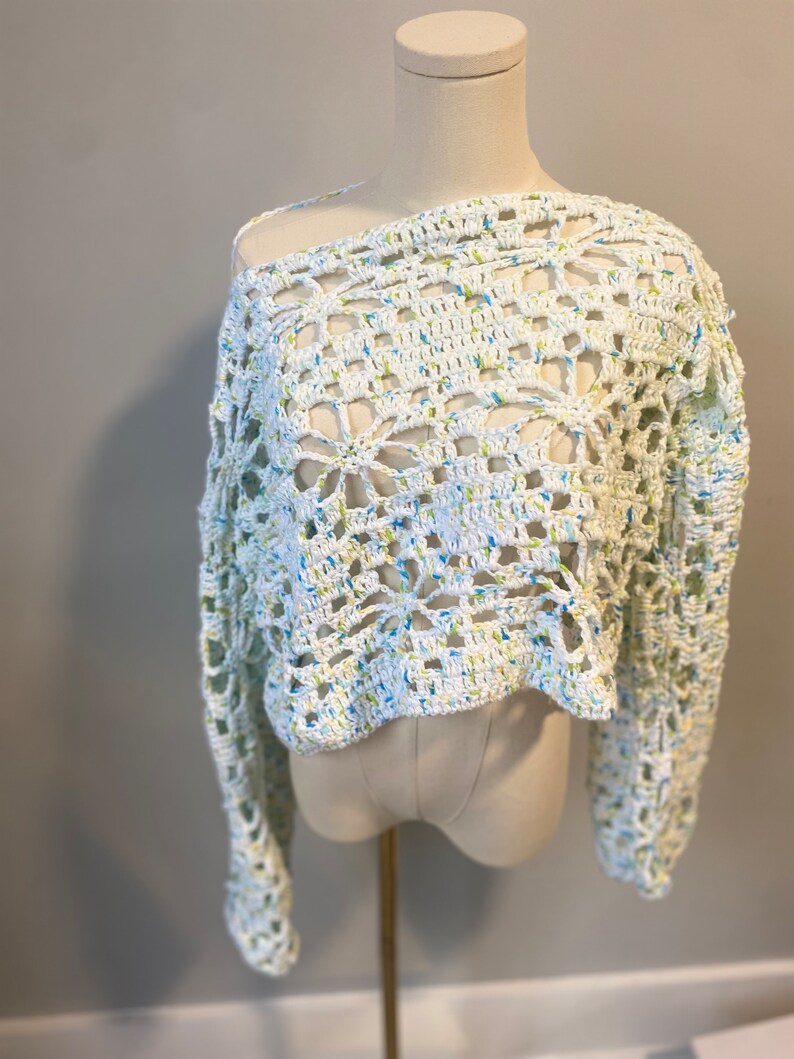 May include: A white, off-the-shoulder crochet top with long sleeves. The open weave design features a floral pattern and is speckled with blue and yellow threads. The top is displayed on a mannequin.
