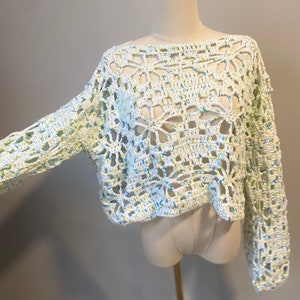 May include: A white crochet top with long sleeves. The open weave design features blue and green flecks throughout. The top is displayed on a mannequin.