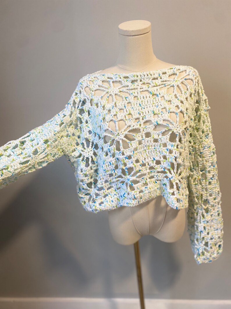 May include: A white crochet top with long sleeves. The open weave design features blue and green flecks throughout. The top is displayed on a mannequin.