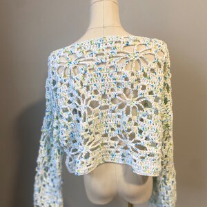 May include: A white crochet top with long sleeves. The openwork design features a floral pattern with blue and yellow accents. The top is displayed on a mannequin.