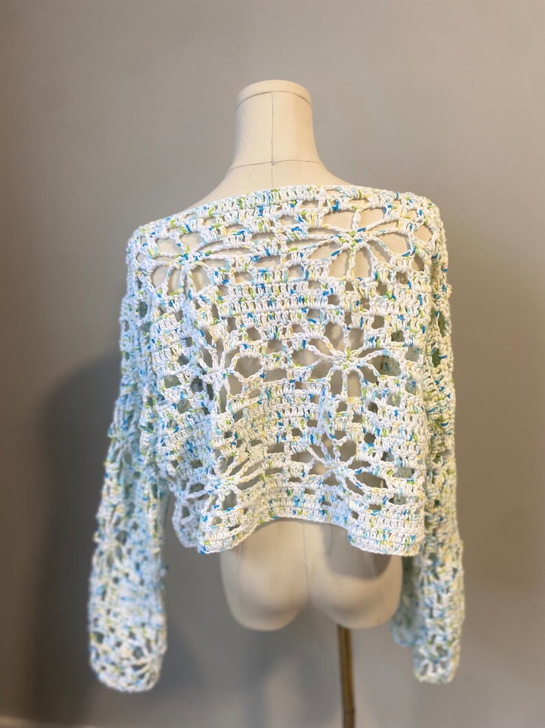 May include: A white crochet top with long sleeves. The openwork design features a floral pattern with blue and yellow accents. The top is displayed on a mannequin.