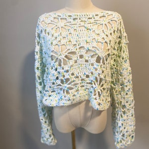 May include: A white, open-weave crochet sweater with long sleeves. The sweater features a floral pattern and is speckled with blue and yellow threads. The garment is displayed on a mannequin against a neutral background.