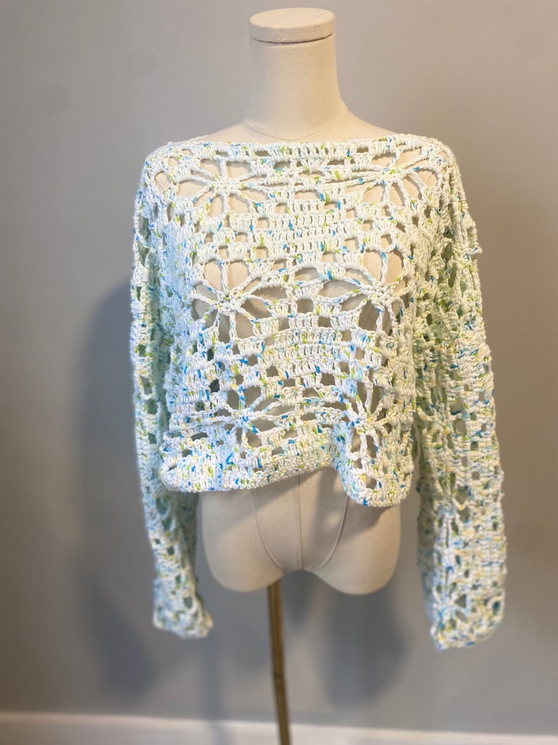 May include: A white, open-weave crochet sweater with long sleeves. The sweater features a floral pattern and is speckled with blue and yellow threads. The garment is displayed on a mannequin against a neutral background.