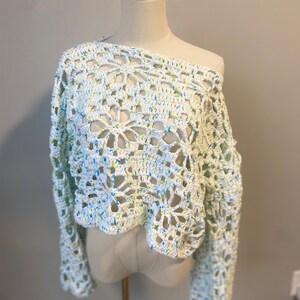 May include: A white, off-the-shoulder crochet sweater with long sleeves. The open weave design features a floral pattern and is speckled with blue and yellow threads. The sweater is displayed on a mannequin.