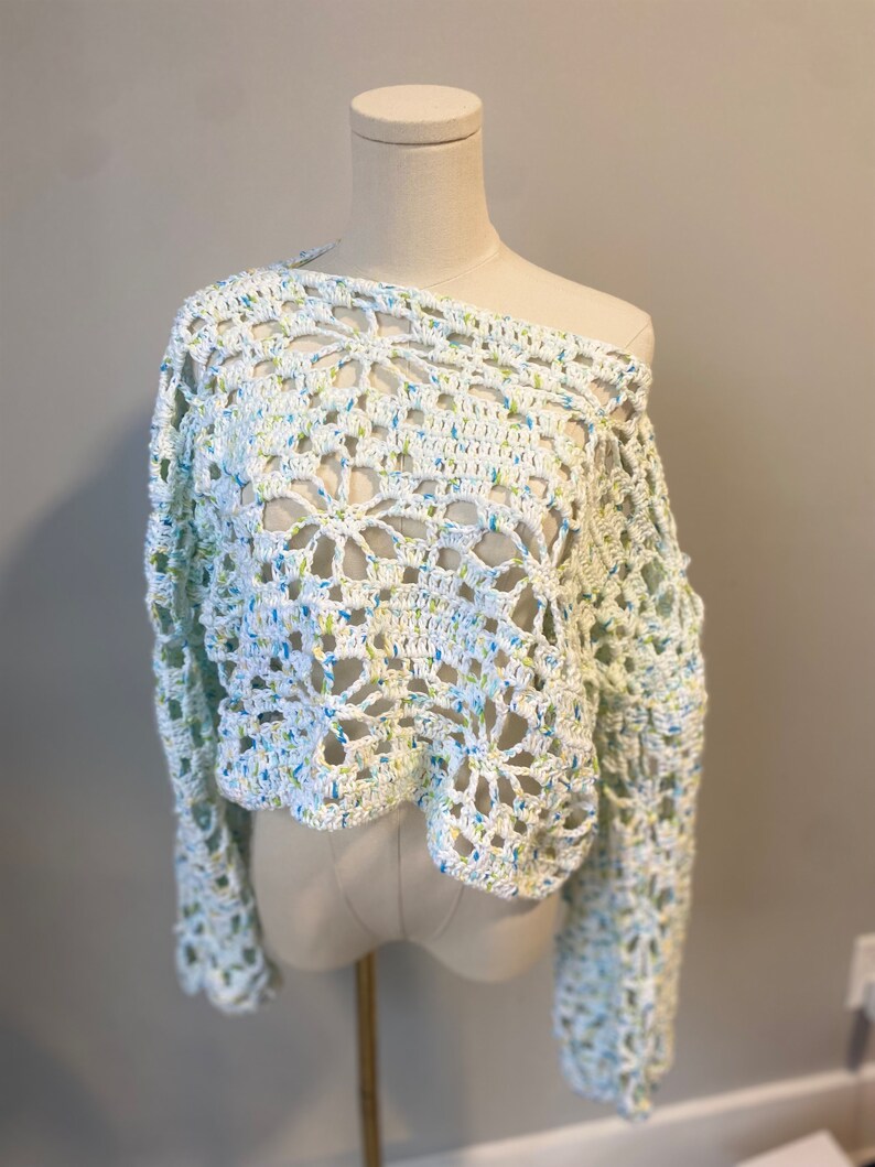 May include: A white, off-the-shoulder crochet sweater with long sleeves. The open weave design features a floral pattern and is speckled with blue and yellow threads. The sweater is displayed on a mannequin.