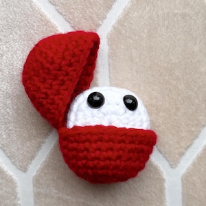 Puede incluir: Un amigurumi blanco de ganchillo con ojos negros asomando de una funda roja de ganchillo.