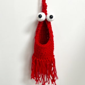 May include: A red crocheted hanging pouch with a large opening at the top and a fringe at the bottom. The pouch has two large white googly eyes with black pupils.