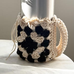 May include: A crocheted black and cream beverage cozy with a handle. The cozy is made with a granny square pattern.