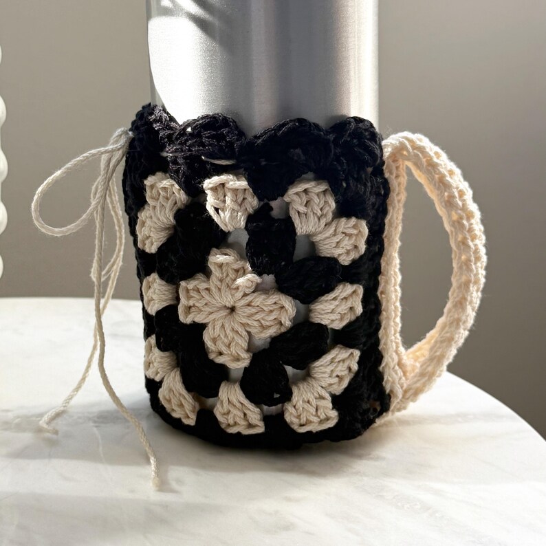May include: A black and cream crocheted mug cozy with a handle. The cozy is made with a granny square pattern.