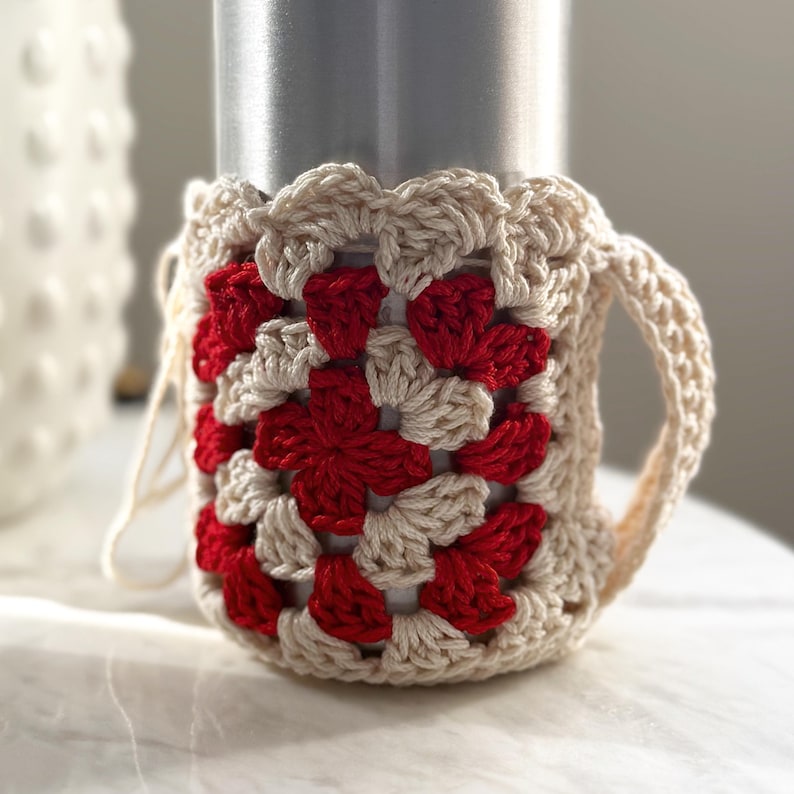 May include: A crocheted cup cozy with a red and white granny square pattern. The cozy is made with a cream colored yarn and has a handle.
