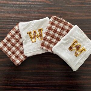 以下が含まれることがあります： 4つの正方形の布製コースター。2つは白で、金と茶色の文字「W」が刺繍されています。他の2つは茶色と白のチェック柄です。コースターは濃い木材の表面に配置されています。