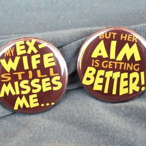 May include: Two brown button pins with yellow text. The first pin says "My ex-wife still misses me..." The second pin says "But her aim is getting better!"