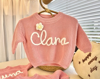 May include: A pink knitted sweater with a white flower and the name "Clara" embroidered on the front.
