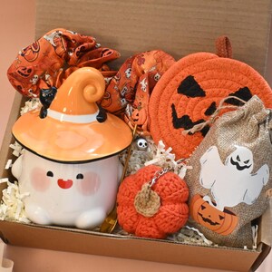 May include: A Halloween-themed gift box filled with festive items. Includes a ceramic ghost mug with a witch hat, a pumpkin-shaped coaster, a burlap bag with a ghost graphic, and a crocheted pumpkin.