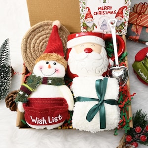May include: A Christmas gift box containing festive items. It features a snowman plush with a red "Wish List" pocket, a Santa mug, a white plush item tied with a green ribbon, and a "Merry Christmas" card. A small Christmas tree and decorative accents complete the arrangement.