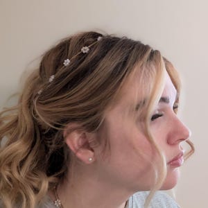May include: A woman with long, wavy brown hair wearing a grey t-shirt and a delicate gold necklace with small white flowers. She is also wearing a gold headband with small white flowers.