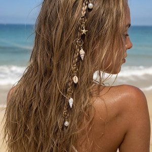 Può includere: Una treccia di capelli decorata con anelli in metallo dorato e ciondoli pendenti, tra cui una perla bianca, una piccola stella marina e varie conchiglie. L'accessorio è intrecciato tra i lunghi capelli castano chiaro e ondulati.