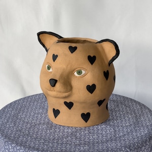 May include: A tan ceramic planter shaped like an animal head with black heart patterns and green eyes.