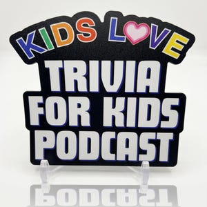以下が含まれることがあります： KIDS LOVE TRIVIA FOR KIDS PODCASTと白い文字で書かれた黒い看板。KIDSという単語はマルチカラーの文字で、LOVEのOの文字はピンクと白のハートに置き換えられています。看板は透明なプラスチック製のスタンドに立てかけられています。