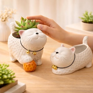 May include: Two white ceramic cat planters with a textured surface. One cat holds an orange ball. Each cat has a black collar with a gold bell. One cat has a succulent plant inside. The planters are on a wooden surface.