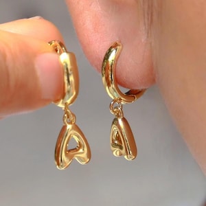 Może przedstawiać: Złote kolczyki koła z wiszącą zawieszką w kształcie litery "A". Kolczyki wykonane są z błyszczącego, odbijającego światło metalu i pokazane są z bliska. Litera "A" ma stylizowany, zaokrąglony kształt.