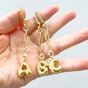Op de afbeelding: Drie goudkleurige initiaalbedels, die "ABC" spellen, hangen aan gouden kettingen en sluitingen. De letters zijn in een afgerond, bubbelachtig lettertype. De achtergrond is licht en neutraal.