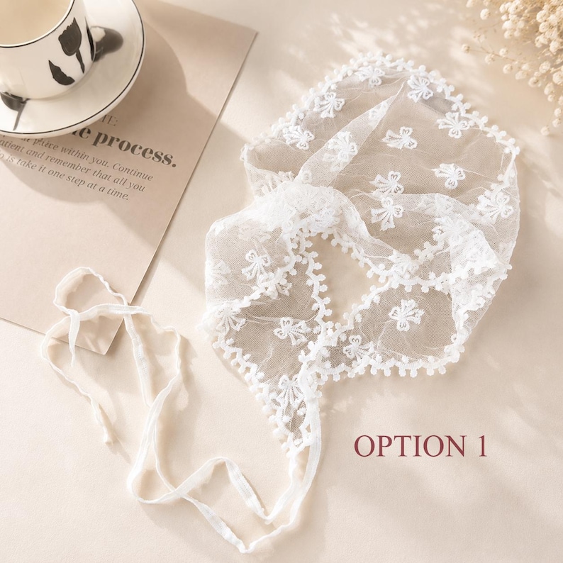 May include: A white, delicate lace head covering with a floral pattern and a ribbon tie. The head covering is labeled "OPTION 1" in a burgundy font. A teacup and saucer with a floral design are in the background.